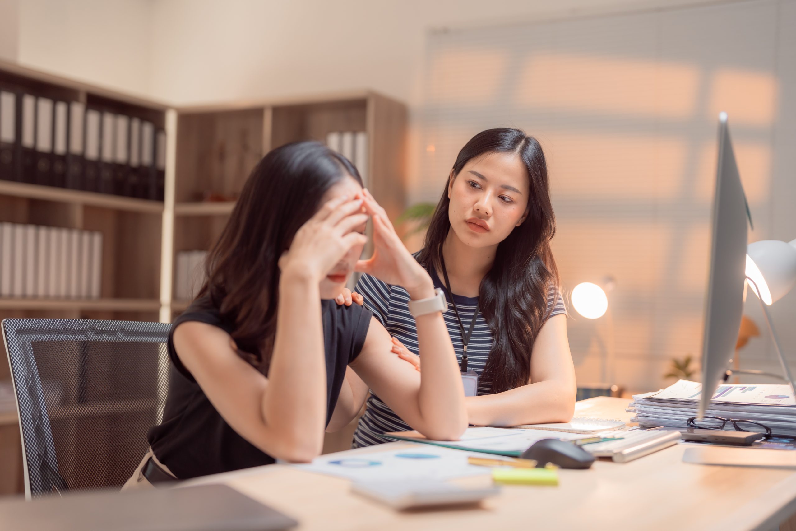 Asian businesswoman is comforting her stressed colleague who is covering her face with her hands, showing concern and support in the workplace