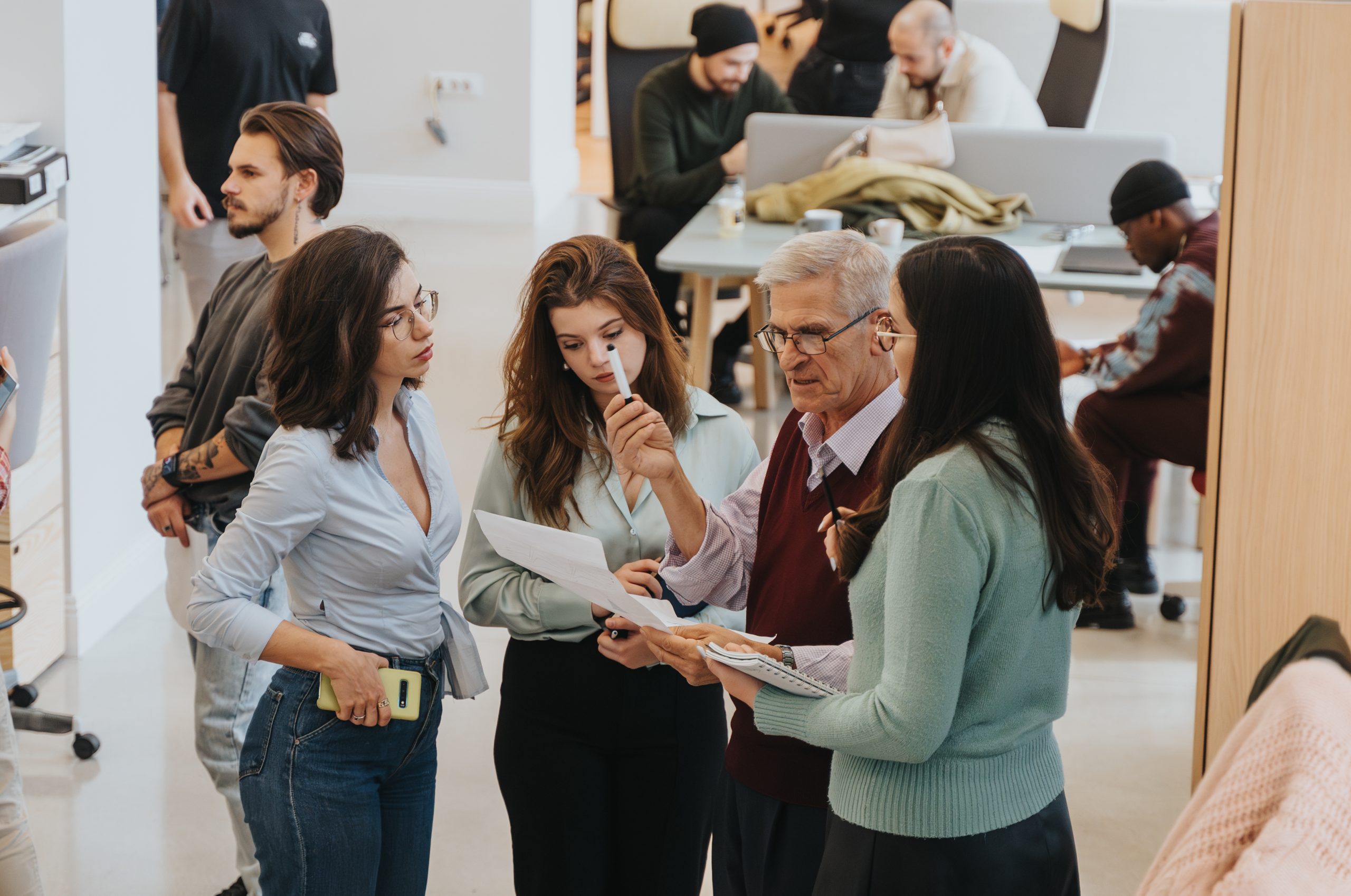 Colleagues collaborate in an office setting, sharing ideas over documents. The focus is on teamwork, communication, and a multi-generational workforce.