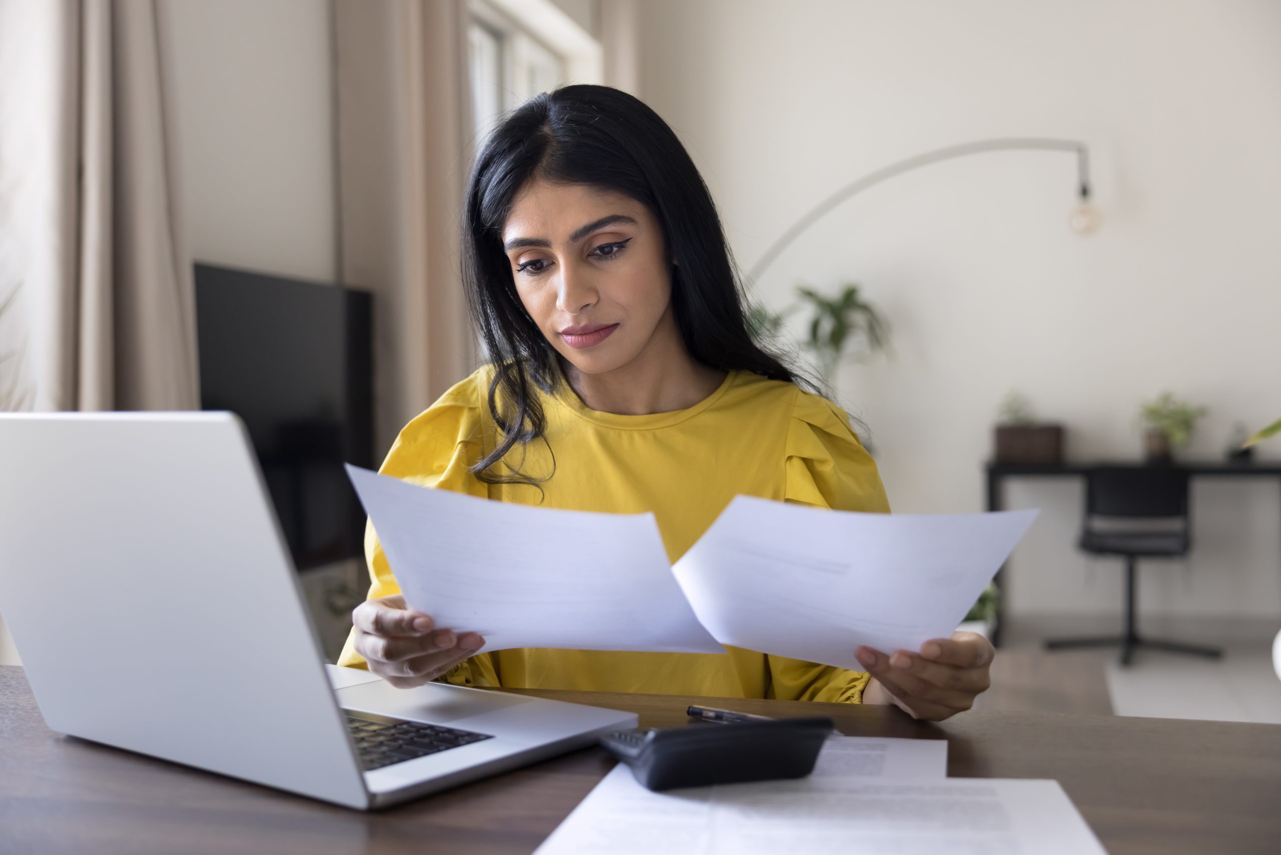 Indian woman engaged in financial work sit at desk with laptop, reviewing documents, checking papers for important details, analyzing contract, read data, looking at bills, invoices, or budget sheets